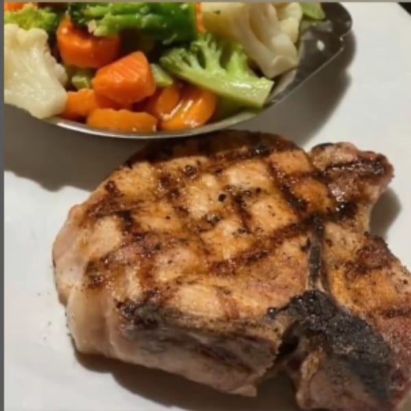 Grilled pork chop with grill marks, served alongside a metal dish of steamed carrots, broccoli, and cauliflower.