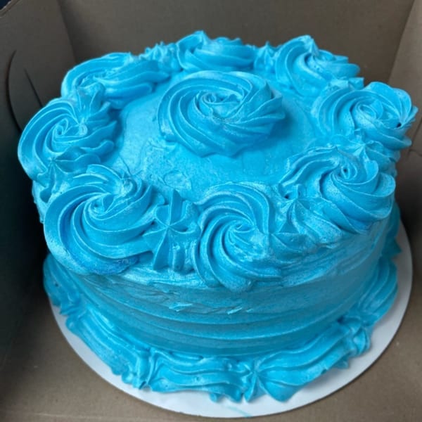 A vibrant blue frosted cake with decorative swirls on top and sides, sitting in a cardboard box.