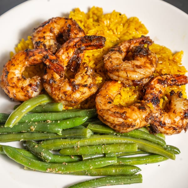 Grilled shrimp with yellow rice and green beans, seasoned with herbs and spices, on a white plate.
