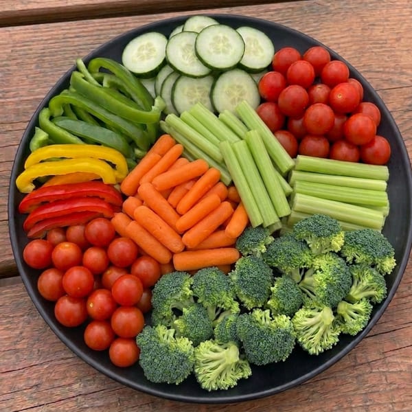 A black platter filled with colorful fresh vegetables, including bell peppers, cucumbers, carrots, celery, cherry tomatoes, and broccoli.