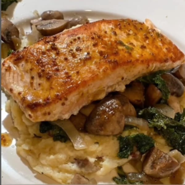 A golden-brown salmon fillet with mustard seeds, served over mashed potatoes, mushrooms, and kale.